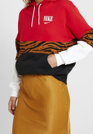 Osoba w czerwonej, białej, czarnej i z tygrysim nadrukiem bluzie Nike z kapturem oraz w musztardowej spódnicy, ręka w kieszeni bluzy, na jednolitym tle.