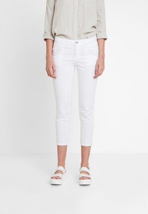 Femme portant une chemise beige à manches longues, un pantalon blanc court et des sandales à plateforme blanches, debout devant un fond blanc uni.