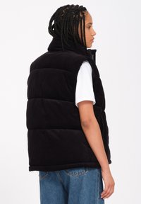 Schwarze Cordweste ohne Ärmel mit hochgelegtem Kragen, ausgestattet mit einem gepolsterten, gesteppten Design. Kombiniert mit einem weißen T-Shirt und blauen Jeans.