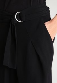 Svarta byxor med en bred midjeband och ett tygbälte, silverfärgad metallring och sidofickor. Slät, matt textur.