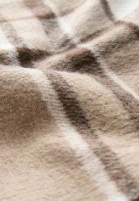 Wełniany koc o miękkiej, puszystej teksturze, w beżowym i brązowym wzorze w kratę, z naprzemiennymi grubymi i cienkimi paskami.