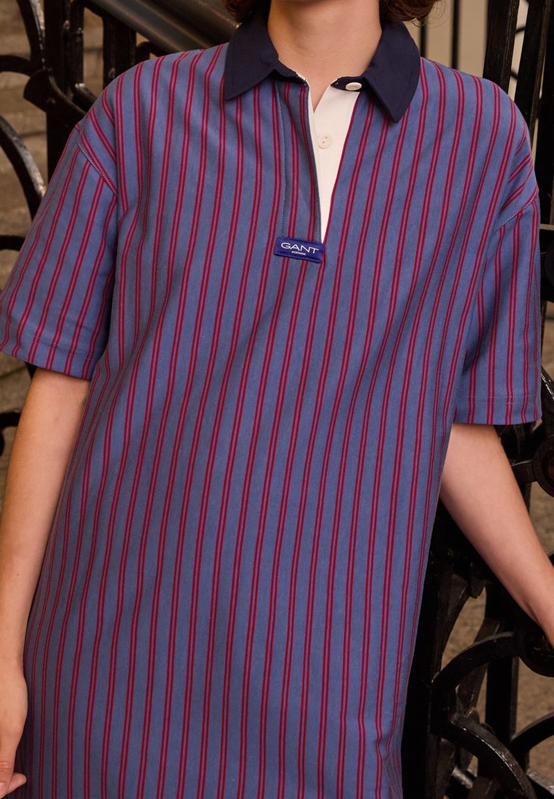 Polo bleu à rayures rouges verticales, avec un col bleu marine et une patte de boutonnage blanche. Logo GANT brodé sur la poitrine. Manches courtes.