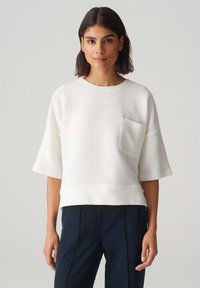 Top bianco a maniche corte con finitura testurizzata, caratterizzato da una tasca frontale e un taglio dritto, abbinato a pantaloni scuri.