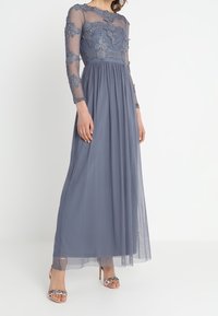 Maxiklänning i skiffergrått med långa genomskinliga ärmar, spetsdekorationer på bysten och en flödande genomskinlig kjol. Buren med dekorerade klackar.