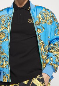 Ljusblå bomberjacka med gyllene barockmönster, med en svart polo skjorta under med broderad logotyp och kontrasterande muddar.