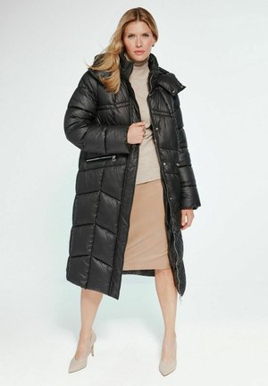 Kobieta ubrana w długą czarną kurtkę puchową, pod którą ma beżowy golf i spódnicę, stoi z jedną ręką trzymając kurtkę przy klatce piersiowej, na nogach ma beżowe szpilki.