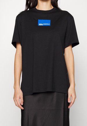 Persona que lleva una camiseta negra de manga corta con un logo rectangular azul de Karl Lagerfeld Jeans en el pecho, combinada con una falda negra.