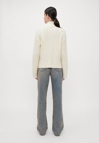 Κρεμ πουλόβερ με πλέξη καμπάνας και ψηλό γιακά, συνδυασμένο με ανοιχτό μπλε φαρδύ παντελόνι, παρουσιάζοντας ένα υφασμάτινο μοτίβο στο πίσω μέρος του πουλόβερ.