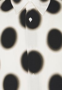 Weißes Hemd mit Knopfleiste, versehen mit großen schwarzen Punkten in einem Verlaufseffekt. Es besitzt einen strukturierten Kragen und Perlmuttknöpfe.