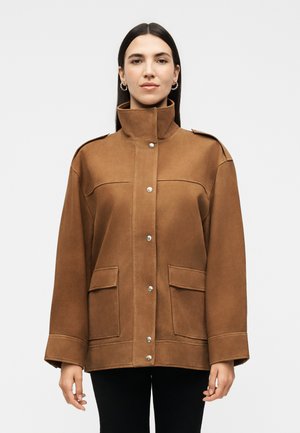 Kobieta z prostymi, ciemnymi włosami nosi brązową kurtkę z wysokim kołnierzem, zatrzaskami i dwoma przednimi kieszeniami z patkami, stojącą na jednolitym tle.