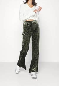 Samtgrüne ausgestellte Hose mit glänzender Textur, kombiniert mit einem weißen Cropped-Pullover und weißen Sneakern. Einfaches Design mit hoher Taille.