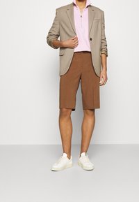 Béžový blazer přes růžovou mikinu na zip s límečkem, v kombinaci s hnědými kalhotami na míru a bílými teniskami. Jsou viditelné strukturované materiály.
