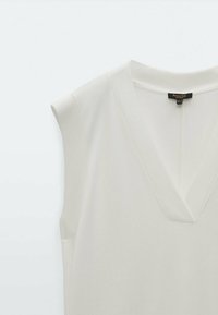 Ärmellose, cremefarbene Bluse mit V-Ausschnitt, glatter Textur und dezentem Stretch. Verfügt über ein Etikett zur Markenidentifikation.