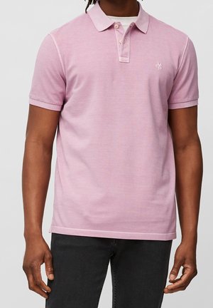 Mann trägt ein hellrosa kurzärmeliges Poloshirt mit Knöpfen und dunkle Hose, steht mit entspannten Armen vor einem schlichten Hintergrund.