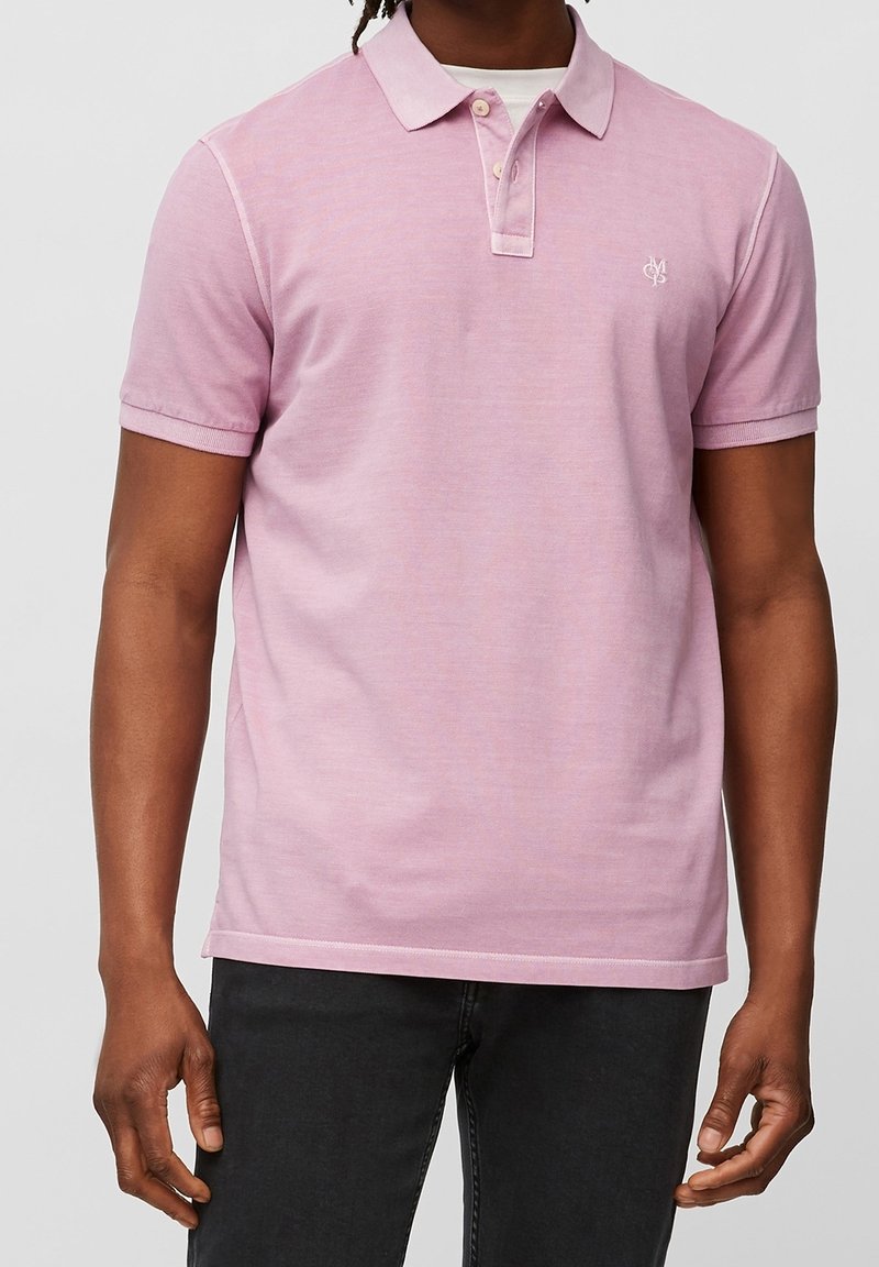 Homme portant un polo rose pâle à manches courtes avec des boutons et un pantalon foncé, debout les bras détendus devant un fond uni.