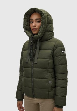 Femme portant une veste matelassée vert foncé avec une capuche relevée et une fermeture éclair à l'avant, debout devant un fond clair uni.