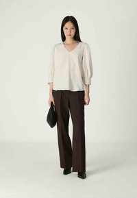 Light beige blouse with puff sleeves and a V-neck, paired with dark brown wide-leg trousers. Model holds a small black handbag.