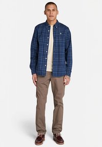 Blå rutig skjorta med knappar, beige t-shirt, bruna byxor och mörkbruna snörskor. Person står mot en enfärgad bakgrund.