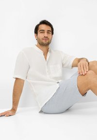 Camicia bianca in lino con colletto corto e tasca sul petto, abbinata a pantaloni corti a righe azzurre. Texture liscia, design casual.
