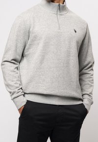 Graues Sweatshirt aus Baumwolle mit Viertel-Reißverschluss und geripptem Kragen. Verfügt über ein kleines Logo auf der Brust und Bündchen an den Ärmeln. Wird über dunklen Hosen getragen.