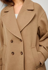 Dubbelzijdige bruine jas van textuurstof, met een brede kraag, twee voorzakken en donkere knopen.