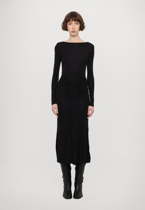 Abito lungo nero con maniche lunghe e scollatura a barca, dettaglio arricciato in vita e gonna fluente. Indossato con stivali neri alti fino al ginocchio.