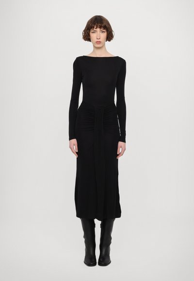 Schwarzes langärmliges Kleid mit einem Bootsausschnitt, Raffung an der Taille und einem fließenden Rock. Getragen mit kniehohen schwarzen Stiefeln.