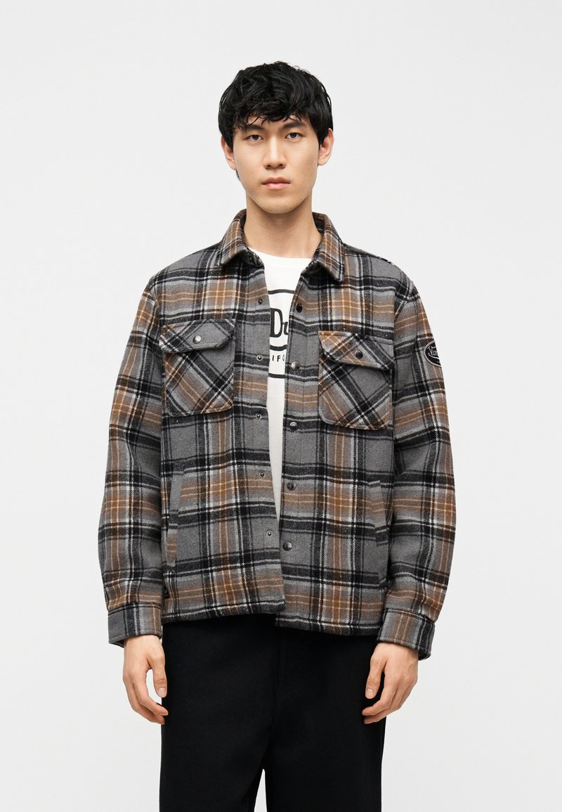 Νέος άνδρας φορώντας γκρι, καφέ και μαύρο καρό μπουφάν πάνω από λευκό μπλουζάκι με μαύρο παντελόνι, στέκεται μπροστά σε απλό φόντο.