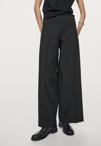 Zwarte wijde broeken met een gladde textuur, hoge taille en een tailored ontwerp. Gecombineerd met zwarte enkellaarsjes, wat een monochrome uitstraling creëert.