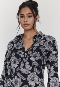 Camicia floreale con sfondo blu navy e motivi di fiori e foglie bianchi. Include un colletto e una scollatura con bottoni. Tessuto morbido.