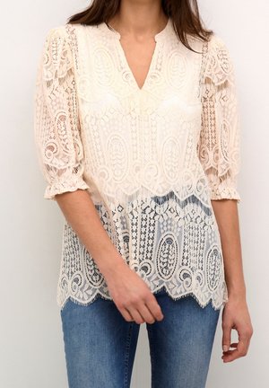 Femme portant un chemisier en dentelle crème à manches trois-quarts et ourlet festonné, associé à un jean bleu, debout devant un fond uni.