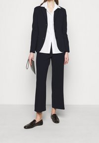 Blazer navy ajustado, camisa blanca de botones, pantalones anchos navy a juego y mocasines marrones oscuros. Cartera en la mano. Texturas suaves.