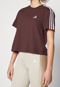 Brun kortärmad t-shirt med vit Adidas-logga och tre vita axelränder. Kombinerad med ljusbeige leggings som har en liten logga.