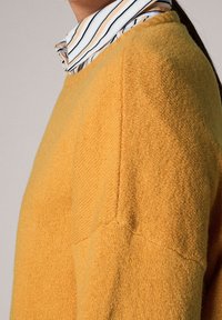 Un pull en maille jaune avec une finition texturée, un col côtelé et des coutures d'épaules tombantes, superposé à une chemise à col rayée.
