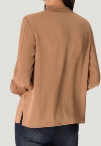 Blouse marron texturée avec un col smocké, des manches longues et des fentes sur les côtés, assortie à un jean foncé.