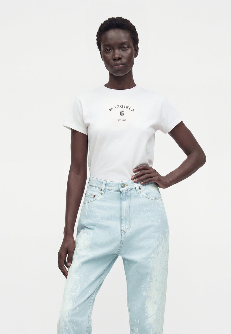 Vrouw met kort krullend haar, gekleed in een wit "Margiela 6" T-shirt en lichtblauwe versleten high-waisted jeans, staand met hand op heup.