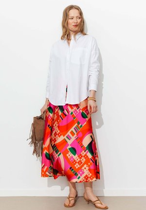 Femme portant une chemise blanche boutonnée et une jupe midi colorée et à motifs, tenant un sac à main marron à franges et portant des sandales.