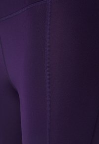 Fialové sportovní legíny s hladkým, pružným materiálem a zesílenými švy. Detailní záběr ukazuje texturu a lesk látky.