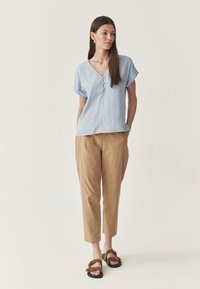 Blusa azzurro chiaro a maniche corte con scollo a V e tre bottoni, abbinata a pantaloni tapered beige e sandali marroni con cinturini.