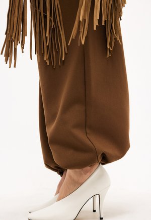 Brązowa tkanina z frędzlami nad luźnymi brązowymi spodniami, stopy w białych szpilkach na jasnym tle.