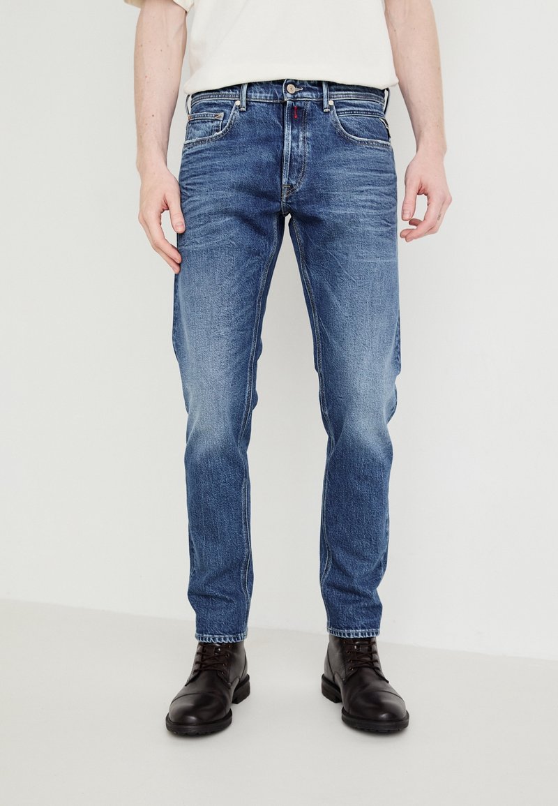 Homme portant un jean slim bleu moyen et des bottes en cuir noires à lacets, debout devant un fond blanc uni.