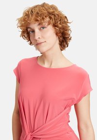 Kortærmet bluse i koralpink med en knude detalje i taljen. Lavet af et blødt, glat stof med en afslappet pasform.