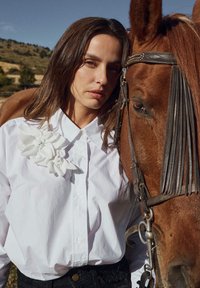 Chemise blanche boutonnée avec de grandes embellissements floraux blancs, associée à un pantalon noir texturé, à côté d'un cheval brun équipé.