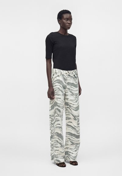 Γυναίκα που φοράει μαύρο τοπ με μανίκια μέχρι τον αγκώνα, φαρδιά ψηλόμεσα παντελόνια με γκρι και λευκό αφηρημένο μοτίβο και μαύρα παπούτσια ανοιχτά μπροστά.