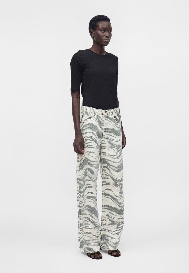 Frau trägt ein schwarzes Top mit Ellbogenärmeln und weit geschnittene, hochtaillierte Hosen mit grau-weißem abstraktem Muster und schwarzen Sandalen mit offenen Zehen.