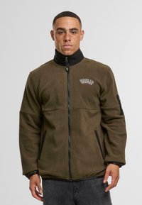 Vihreä fleece-takki korkealla kauluksella, mustat yksityiskohdat vetoketjussa ja taskuissa sekä brodeerattu "UNFAIR" -logo rinnassa.