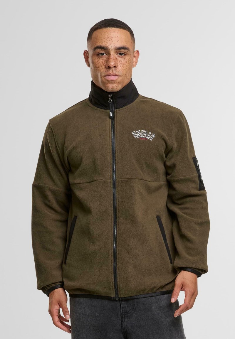Vihreä fleece-takki korkealla kauluksella, mustat yksityiskohdat vetoketjussa ja taskuissa sekä brodeerattu "UNFAIR" -logo rinnassa.