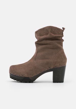 Bota de ante marrón hasta media caña con parte superior caída, adornos con tachuelas, suela con taco y tacón bloque sobre un fondo claro y liso.