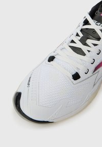 Witte sportschoen met zwarte en roze accenten, witte veters, mesh textuur en "FLEXSHIELD" gedrukt nabij de teen.