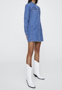Robe-chemise en denim à manches longues, fermeture à boutons sur le devant, deux poches poitrine, dans une teinte bleu clair, associée à des bottes western blanches.
